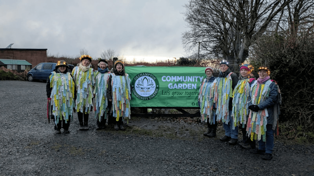a picture of 8 people standing in front of a Okehampton community garden banner, with fairy lights on and outfits made from recycled fabric for the celebration of Wassail.