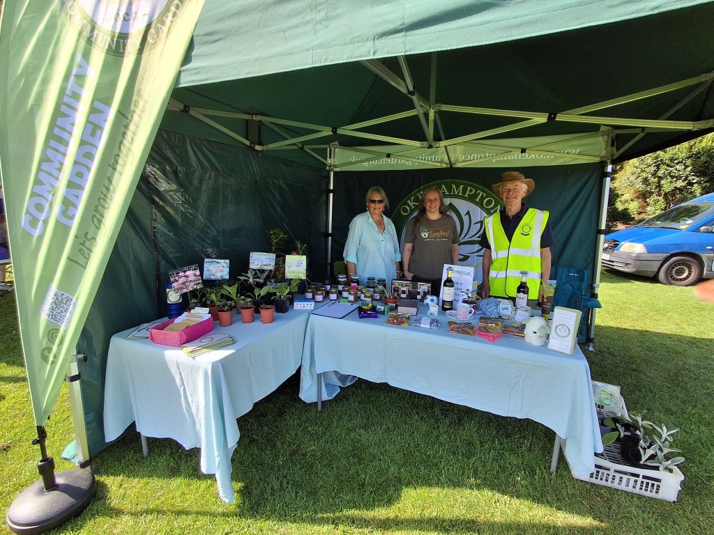 Three volunteers stand behind a community garden stall beneath a green gazebo at an outdoor event. Tables display plants, preserves, information leaflets, and fundraising items, promoting the work of Okehampton Community Garden.