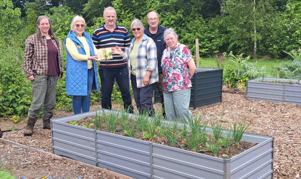 Six people stand together in a community garden beside raised vegetable beds, holding a plaque or award. The garden features woodchip paths, thriving greenery, and mature trees in the background, highlighting a productive and welcoming outdoor growing space.