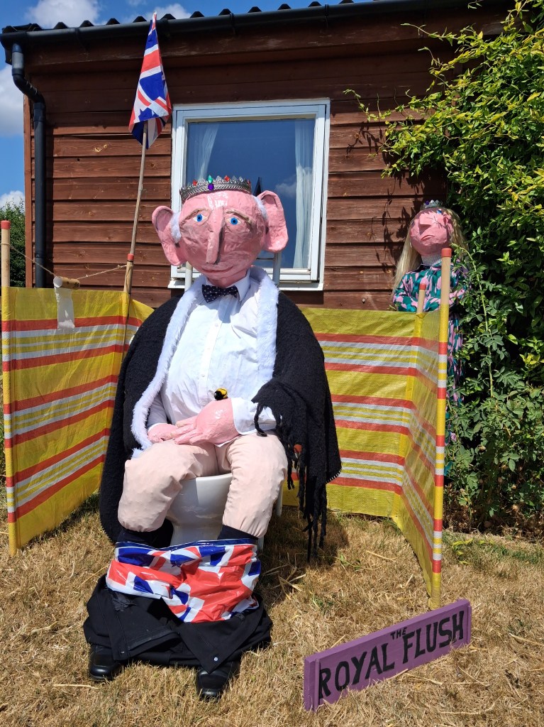 Scarecrow competition entry titled 'The Royal Flush' featuring a humorous royal figure wearing a crown and ceremonial robe, seated on a toilet behind a striped windbreak. A Union Jack flag flies above, with another crowned figure peeking from behind a hedge.