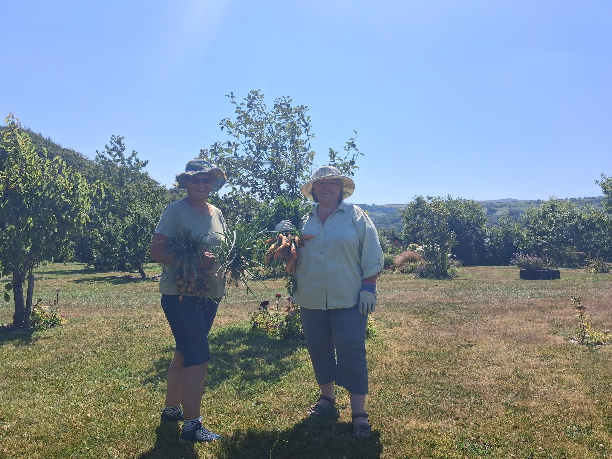 Two volunteers stand in Okehampton Community Garden holding freshly harvested carrots and onions, smiling in the sunshine with trees and countryside in the background.